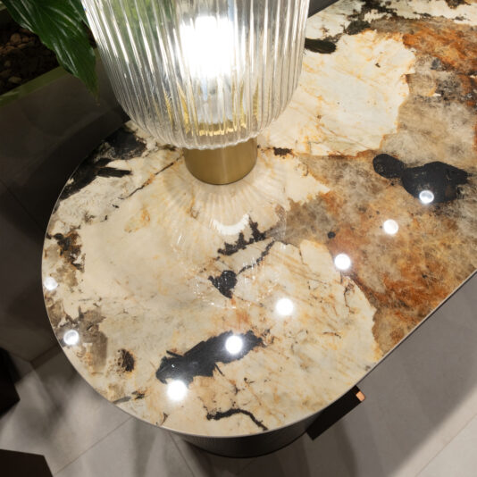 A close-up of a polished stone tabletop with beige, black, and rust hues reflects light from a ribbed glass lamp with a brass base, set beside the Contemporary Hammered Oak Veneer Sideboard.