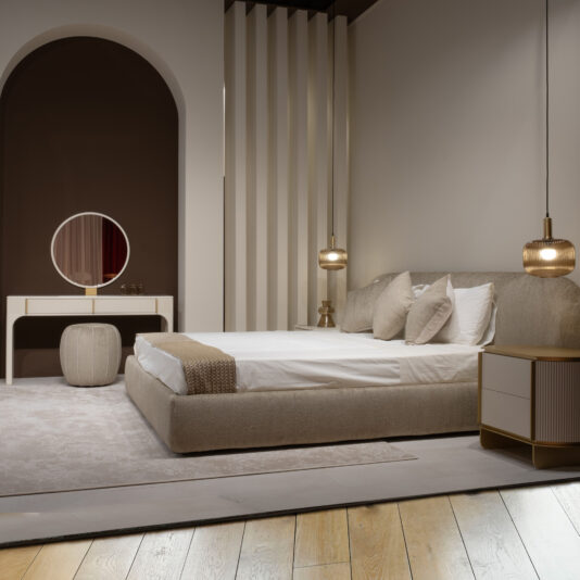 Modern bedroom featuring a beige upholstered bed, cream bedding, two pendant lights, a round mirror on the Contemporary Lacquered Dressing Table, a pouffe, bedside table, soft neutral walls, and light wood flooring.