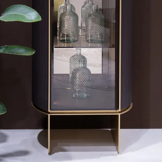 The Contemporary Lacquered Narrow Display Cabinet with gold trim features three textured glass bottles on two shelves. A green plant leaf sits to the left, against a brown wall and light flooring.