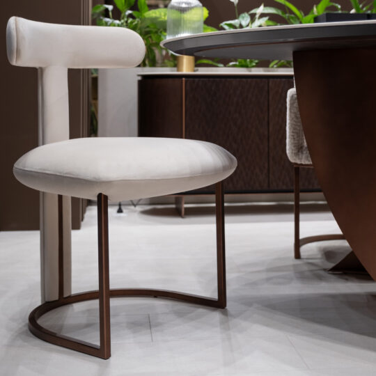 The Modern T Backed Dining Chair (Copy), featuring a curved metal base and plush round white seat and backrest, is placed by a dining table with green plants and a wooden sideboard in the background.