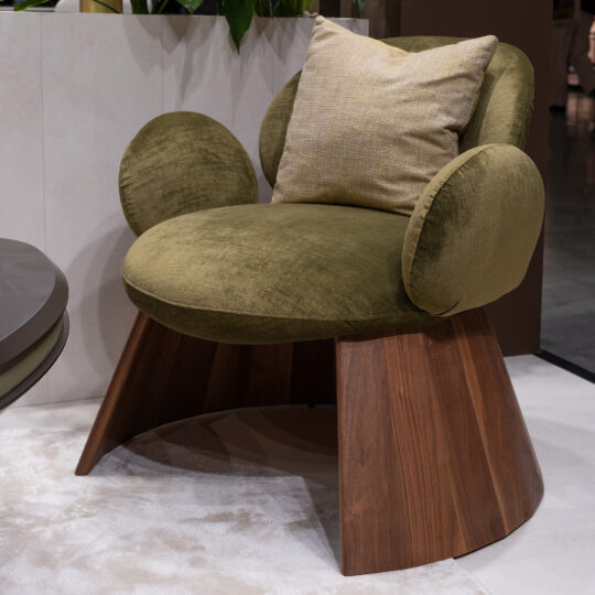 The Contemporary Walnut Statement Armchair in green velvet has rounded armrests and backrest, paired with a light beige cushion and set on a wooden angled base atop a light carpet.
