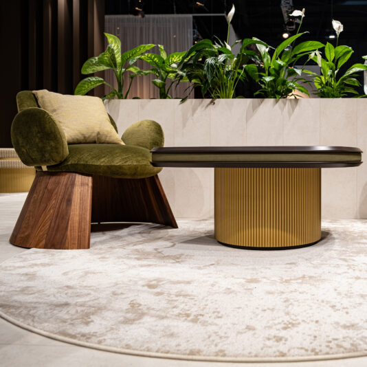 Contemporary Walnut Statement Armchair A modern lounge area with the Contemporary Walnut Statement Armchair (green cushions), a round gold-base coffee table, beige rug, and lush green plants in the background.