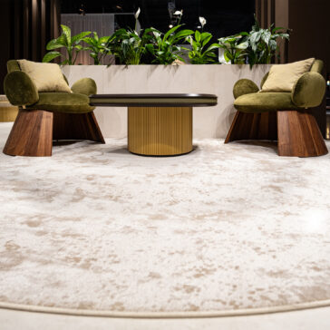 Two Contemporary Walnut Statement Armchairs with green cushions face each other across a round table with a gold textured base on a light patterned rug, whilst lush green plants decorate the background.