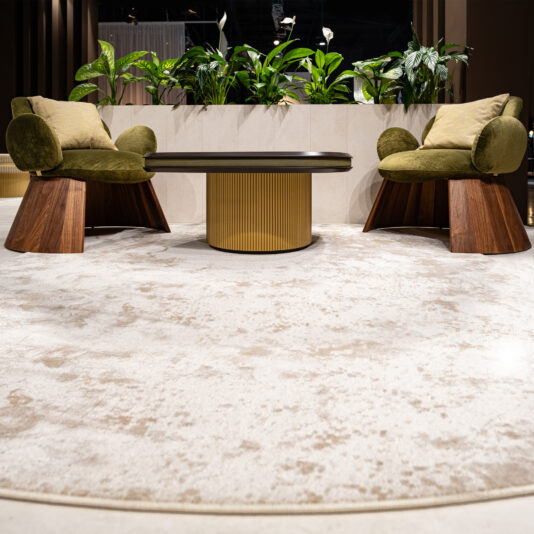 Contemporary Walnut Statement Armchair Two Contemporary Walnut Statement Armchairs with green cushions face each other across a round table with a gold textured base on a light patterned rug, whilst lush green plants decorate the background.