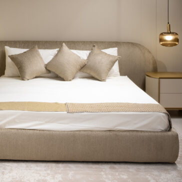 A modern bedroom showcases the Modern Curved Upholstered Bed with four beige scatter cushions, a white sheet, beige bed runner, round bedside table, and a hanging glass pendant light for a neutral and minimalist look.