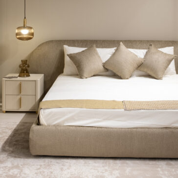 A calm, minimalist bedroom features the Modern Curved Upholstered Bed, three beige scatter cushions, white sheets, a matching bedside table, a small lamp, and a pendant light—all in neutral tones.