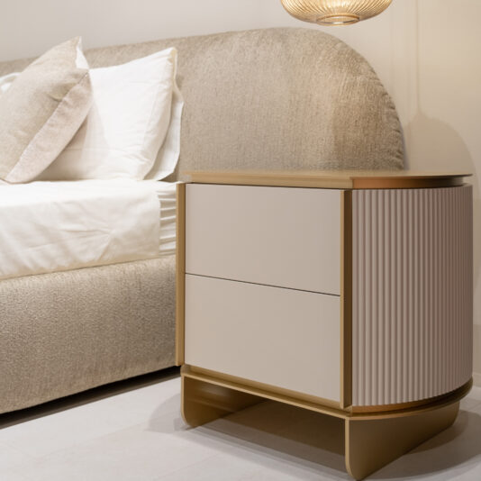 Modern Ribbed Bedside Cabinet The Modern Ribbed Bedside Cabinet with two drawers stands next to a textured beige bed with white bedding and pillows, illuminated by a hanging light in a softly lit, minimalist bedroom.