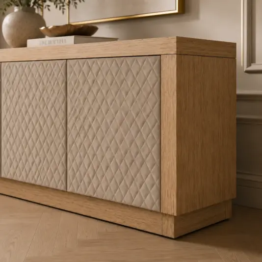 Light wood sideboard with beige, diamond-quilted doors stands on a herringbone wood floor. A decorative book and plant rest on top. The setting appears modern and elegant with soft lighting.