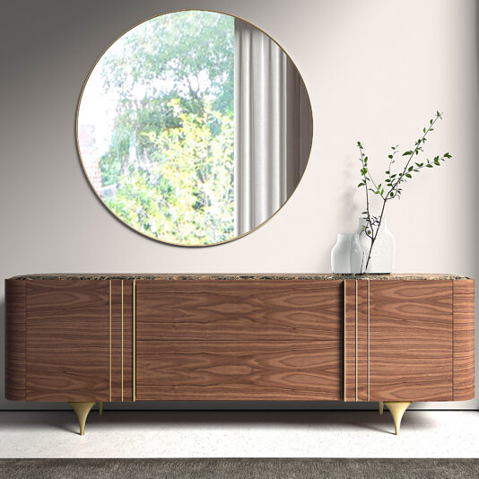 The Contemporary Walnut Veneer Sideboard with gold accents Is the perfect example of the new warm modernism trend for 2027. It stands beneath a large round mirror. A white vase holding a leafy branch rests on top, whilst the mirror reflects nearby greenery and a window.