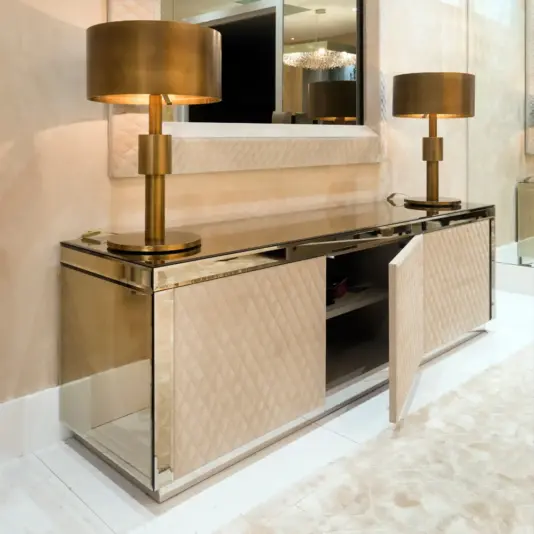 A modern mirrored sideboard with a quilted beige finish stands against a wall, topped with two gold table lamps and a large mirror above. One sideboard door is slightly ajar, revealing shelving inside.